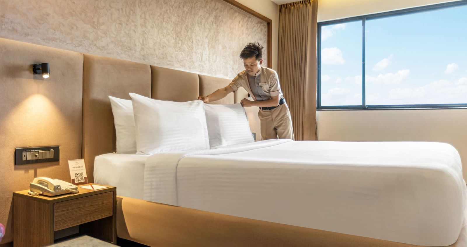 A person arranging pillows on a large bed in a room at Mango Hill Central, Chennai. There is a large window on the right.