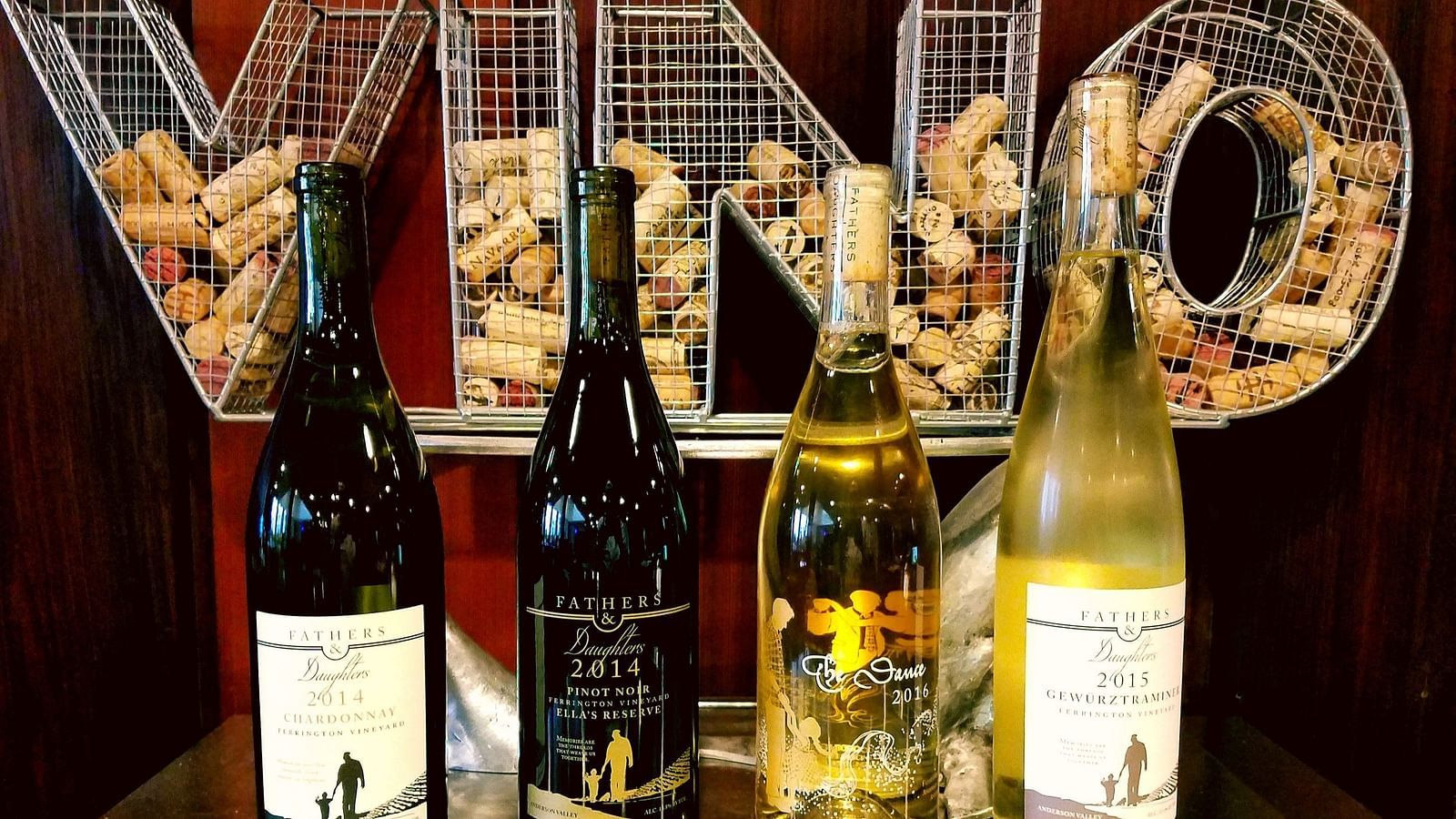 Four bottles stand in a row, behind them wire letters spelling VINO against a dark backdrop..