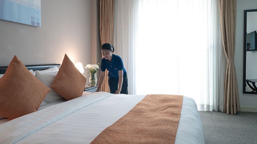 A staff member making a bed, standing beside a large white bed with orange decorative pillows and a matching bed runner near a window with sheer curtains | Central Mansions, Phnom Penh