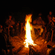 A group of people sitting in a circle around a bright, blazing campfire in the dark of night.