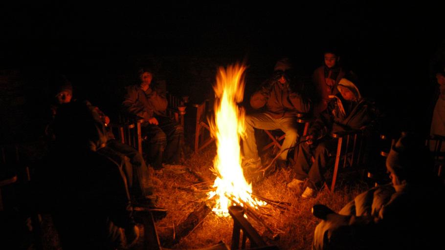 A group of people sitting in a circle around a bright, blazing campfire in the dark of night.