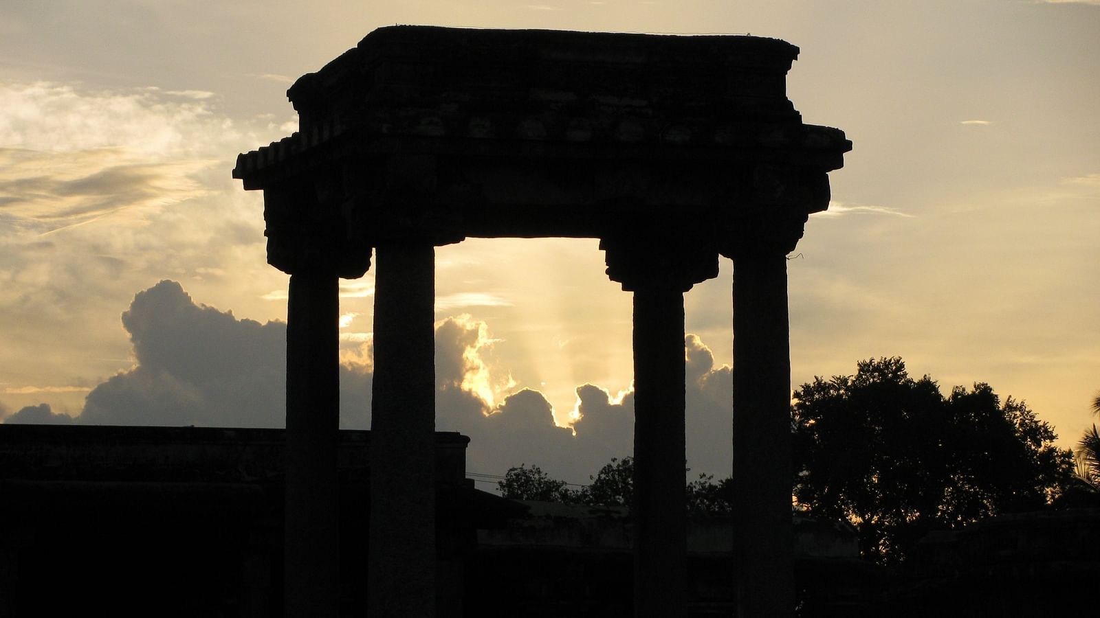 An overview of ancient columns in Belur with the sun's rays behind the clouds in the background.