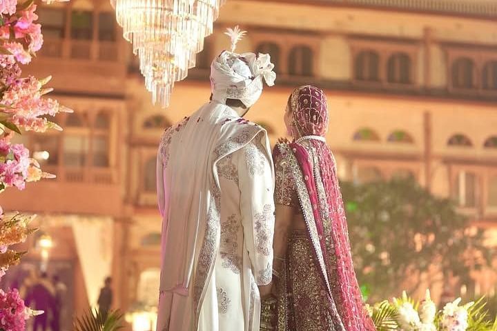 The bride and groom stand hand in hand under a chandelier, surrounded by flowers, with a Heritage Palace Resort in the backdrop - Umaid Palace - A Lakeside Heritage Palace Resort