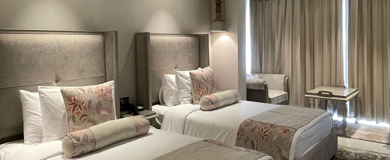 A spacious Premium Twin Room at The Evren, Vagator, with two single beds, each with a plush headboard and decorative pillows. The room features a patterned rug and a seating area near the window.