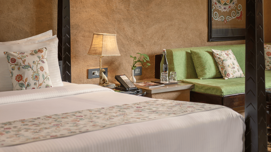 Bedroom interior of Elegant Suite featuring a dark wooden bed, earth-plastered wall with framed traditional art, and a daybed seating area at AamaGhati Wildlife Resort.