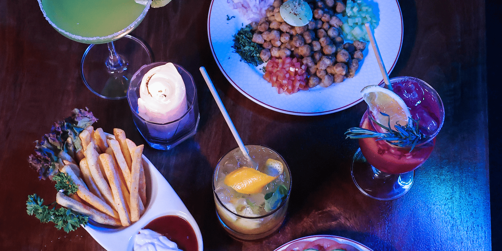 Overhead shot of vibrant food dishes and drinks on a dark wooden table, illuminated by blue and red lighting at Hotel Hukam's Lalit Mahal.
