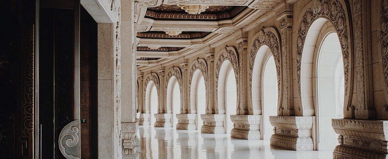 A luxurious hallway with arched windows, columns, and a polished marble floor, evoking a sense of grandeur and opulence.