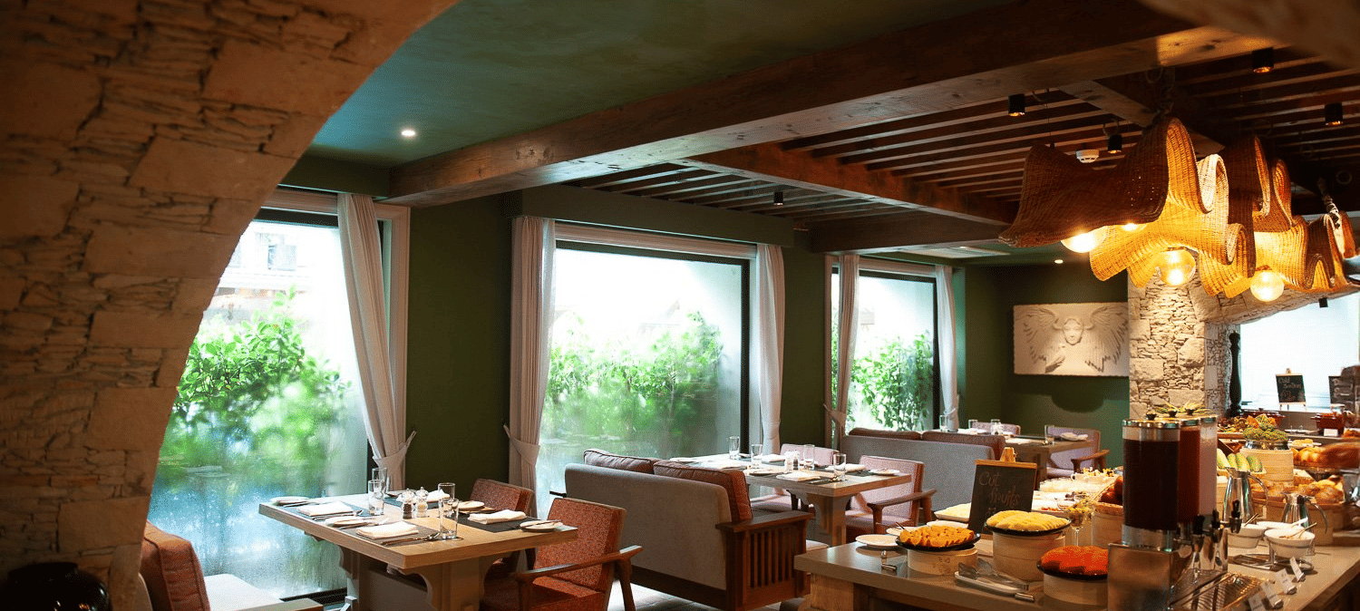A wide-angle view of the dining area at the Mila Restaurant at The Evren, Vagator, from the corner of the stone archway featuring wooden tables, comfortable chairs and banquettes, and a buffet station with a variety of food.