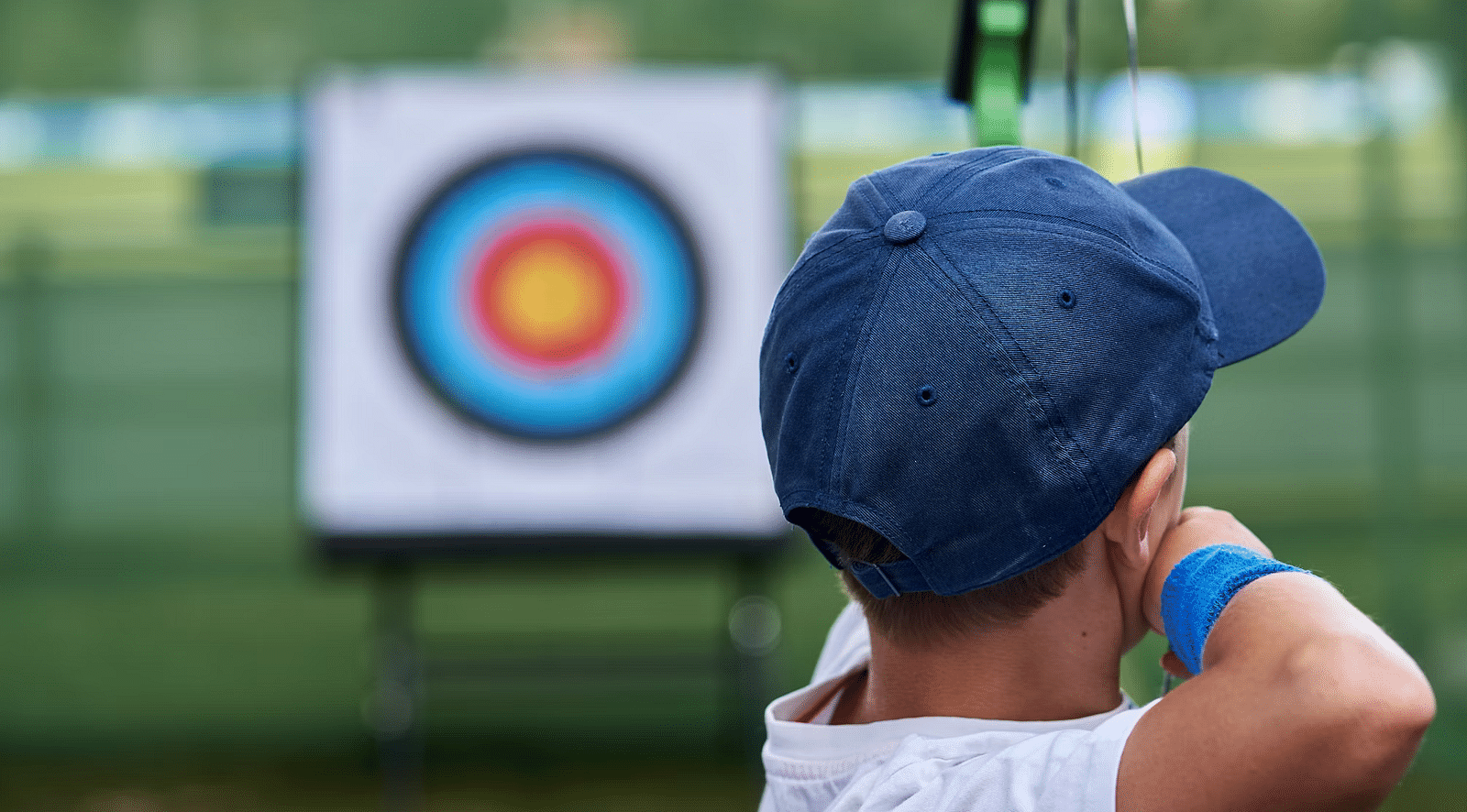 A child wearing a blue cap aims a bow and arrow at a target during archery practice at Nirantara Resort & Spa.