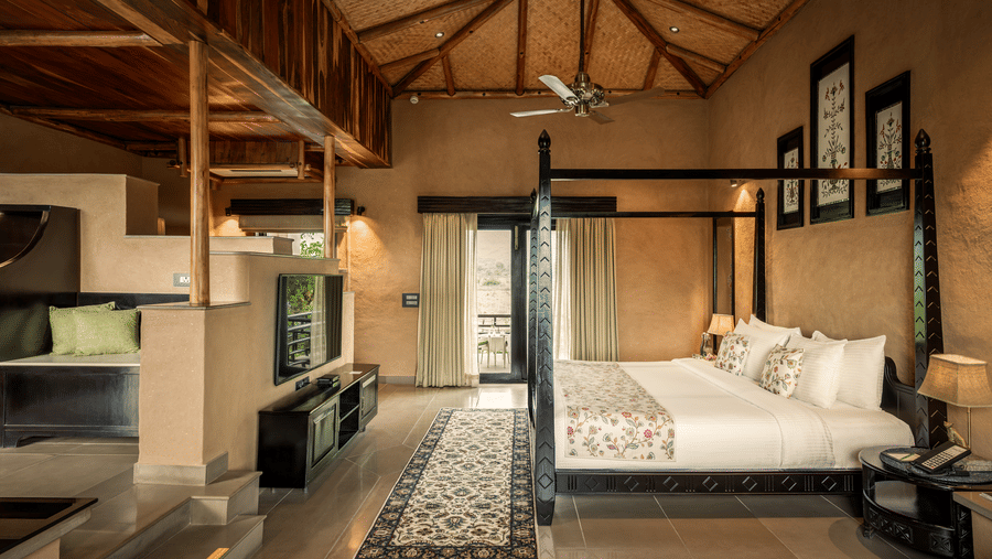 The spacious Elegant Suite at AamaGhati Wildlife Resort featuring a thatched bamboo ceiling, dark wooden bed, split-level living space with a TV console, and access to a outdoor balcony.