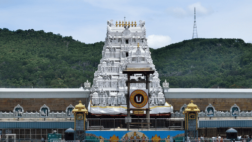 A wide shot of a white, multi-tiered Tirumala Venkateswara Temple tower with golden accents, situated in a courtyard with green hills and a clear sky in the background.