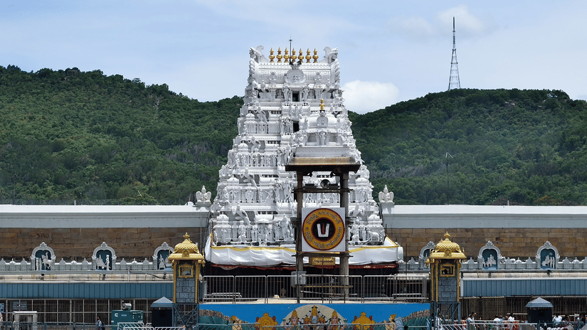 A wide shot of a white, multi-tiered Tirumala Venkateswara Temple tower with golden accents, situated in a courtyard with green hills and a clear sky in the background.