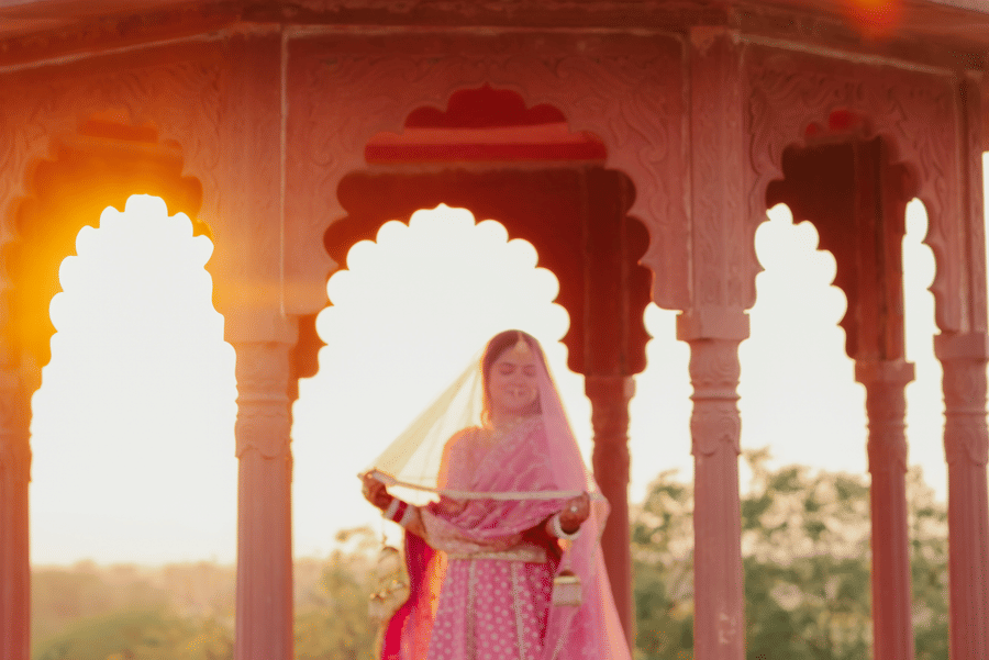A bride in a pink lehenga stands elegantly under a traditional pink stone dome chhatri at Umaid Palace during sunset.