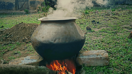 Large metal pot boiling over a wood-fired stove outdoors, with steam rising.