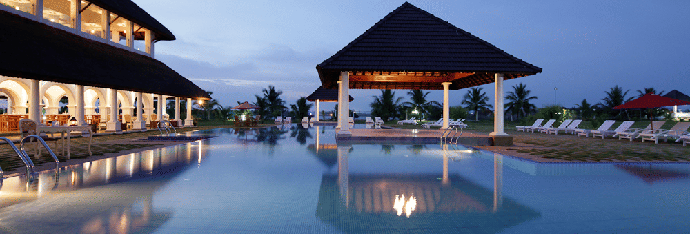 An outdoor swimming pool reflecting a lit pavilion and resort building under a twilight sky with cocktails in the foreground | Le Pondy