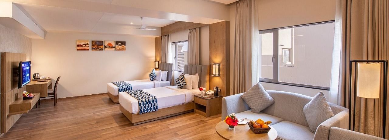 A spacious hotel suite featuring two single beds with blue and white accents, a comfortable seating area with a sofa and armchairs around a small table with fruit, a work desk with a chair, and a television, all on a wooden floor at Lords Inn Motikhavdi