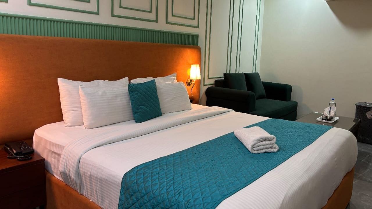 A well-lit room with a double bed dressed in white and blue, and a dark armchair - Asian Suites IFFCO Metro, Gurgaon