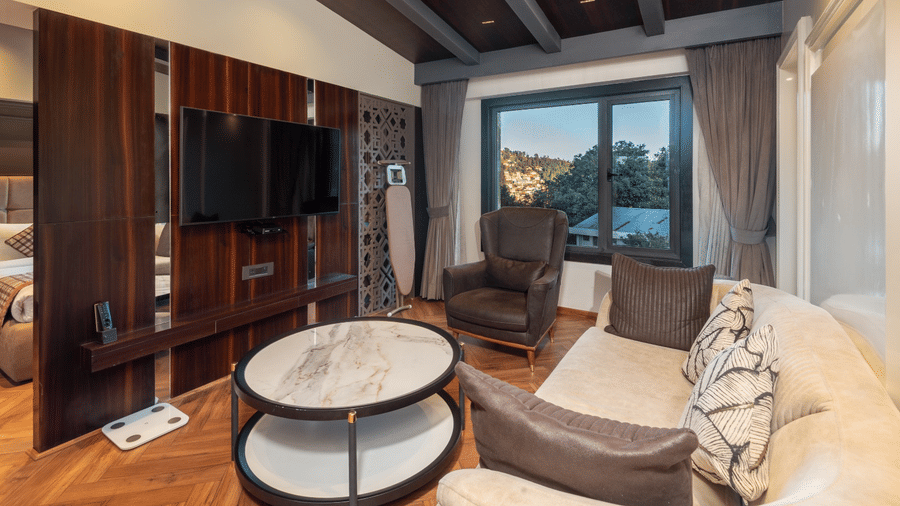A seating area with a sofa, an armchair, and a wall-mounted TV on a dark wood panel next to a large window | Lawrence Solitaire Resort, Nainital