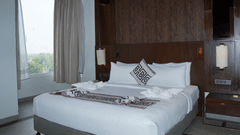 A king size bed with white linens and decorative pillows sits near a large window at Ananta Express, Jaunpur.