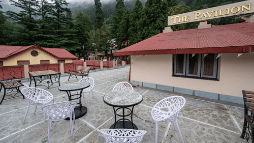 Outdoor terrace at The Pavilion, Nainital with white plastic chairs, round tables, and dramatic misty mountain views through surrounding pine trees.