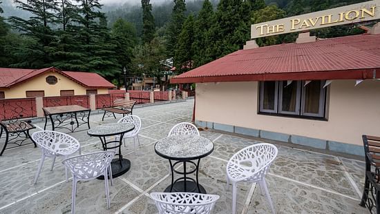 Outdoor terrace at The Pavilion, Nainital with white plastic chairs, round tables, and dramatic misty mountain views through surrounding pine trees.