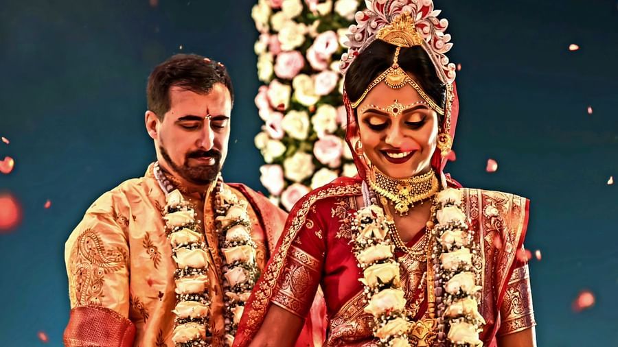 A newlywed Indian couple in traditional attire stands together, with the bride looking down and the groom looking at her, against a softly lit background