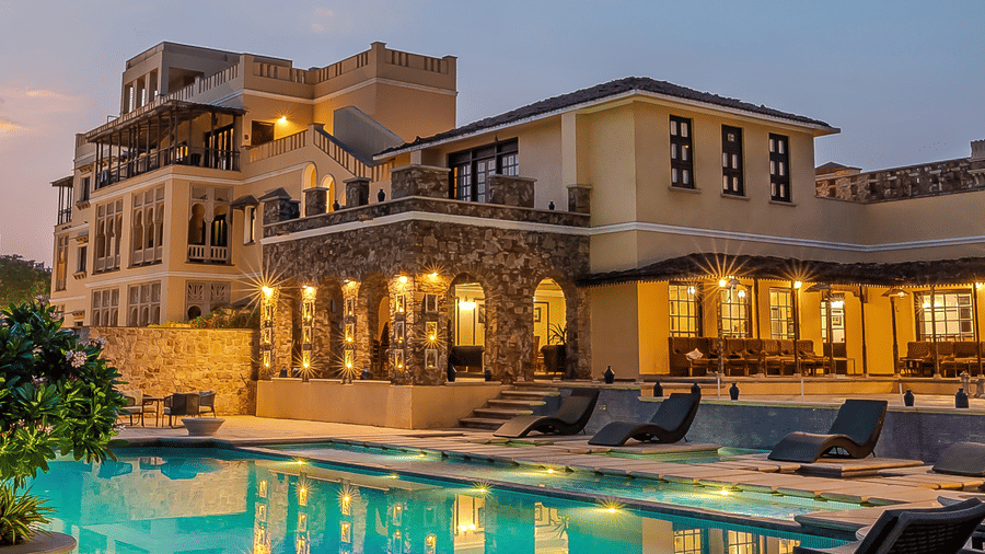 Facade image of Juna Mahal in Ranthambore, Rajasthan featuring a well lit multi storey building, and a swimming pool surrounded by lounger beds and flower pots.