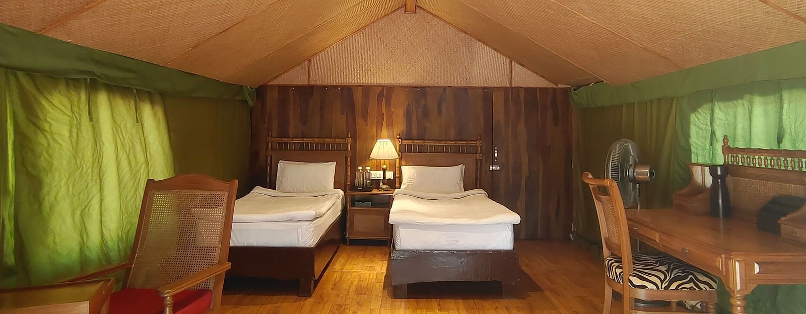 A zoomed out image featuring a cozy room with two beds inside and wooden flooring, lights lit and a lamp next to the beds.