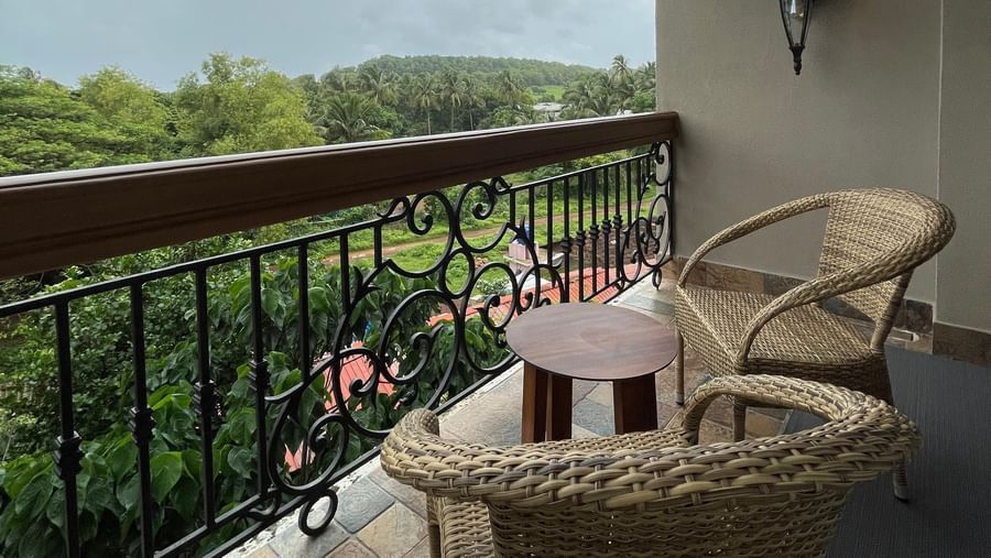 A serene balcony at The Evren, Vagator, showing two wicker chairs and a small table. The balcony overlooks a lush, green landscape with palm trees and fields under a cloudy sky.