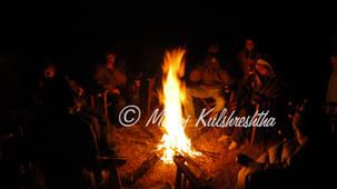 A bright campfire burning at night surrounded by blurred silhouettes of people gathered around.