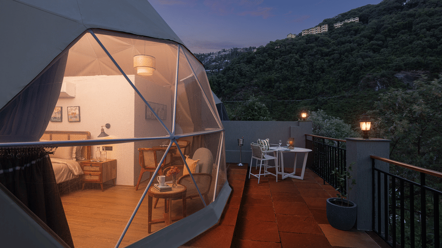 Dusk view of a luxury glamping geodesic dome tent with a clear panoramic window showing a cozy lit bedroom interior.