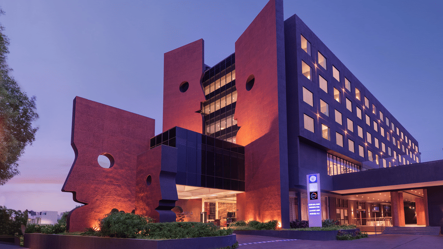 Facade view of Essentia Luxury Hotel, Indore, featuring the illuminated multi-storey building under the blue sky with a large open area in front of the building.