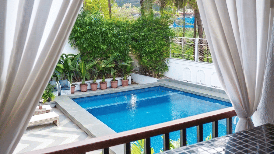 White curtains framed around a window overlooking a swimming pool, outdoor seating, and tropical palm trees at Bougainvillea, Alibaug.
