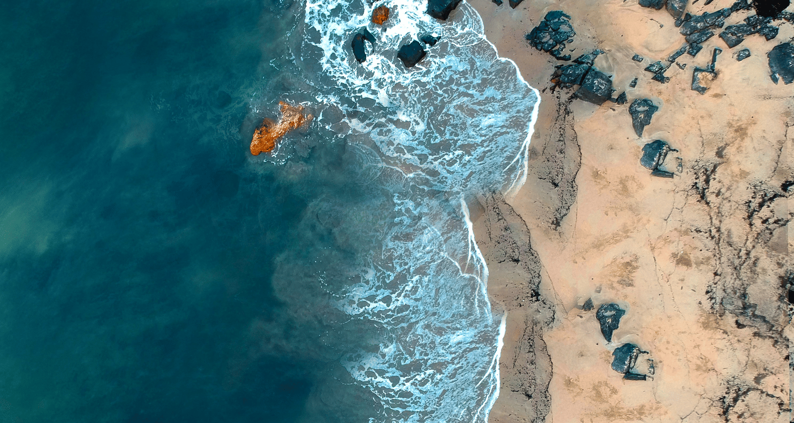 An aerial view of a curved coastline with turquoise water meeting a sandy shore scattered with small rocks.