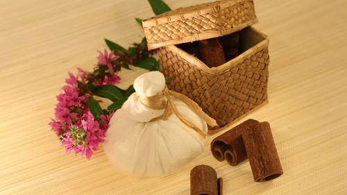 A woven box holds cinnamon sticks next to a fabric herbal compress and flowers on a wooden surface