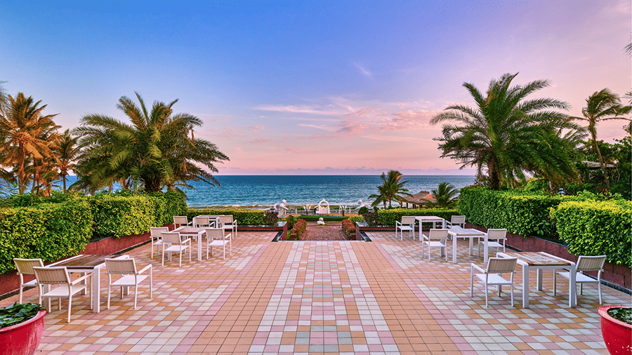 Outdoor terrace dining area with tables set on a tiled patio overlooking palm trees and the ocean at MAYFAIR Palm Beach Resort, Gopalpur-on-Sea.