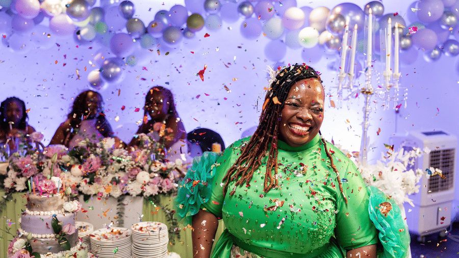 Woman smiling during a wedding celebration with balloons, flowers, and a decorated cake table in the background.
