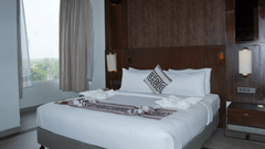 A king size bed with white linens and decorative pillows sits near a large window at Ananta Express, Jaunpur.