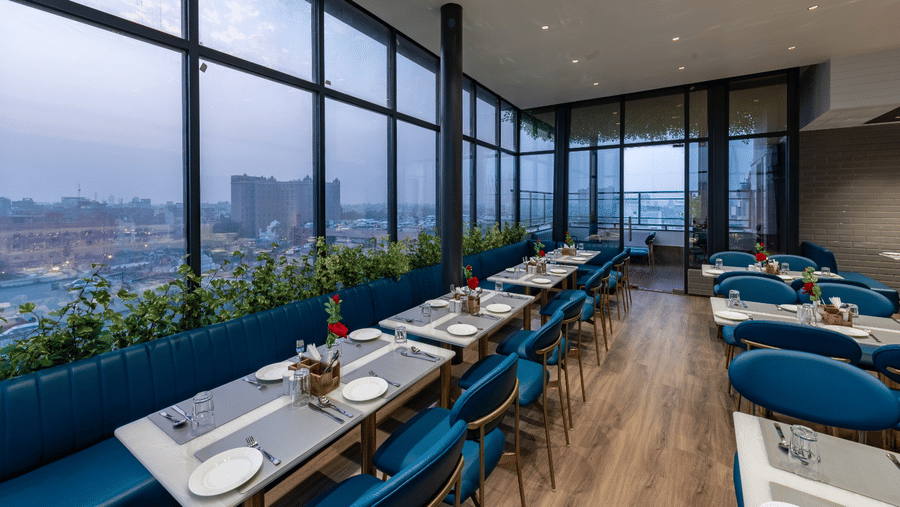 A restaurant with multiple tables, chairs, windows, and an outside views at DLS Hotels Amritsar.