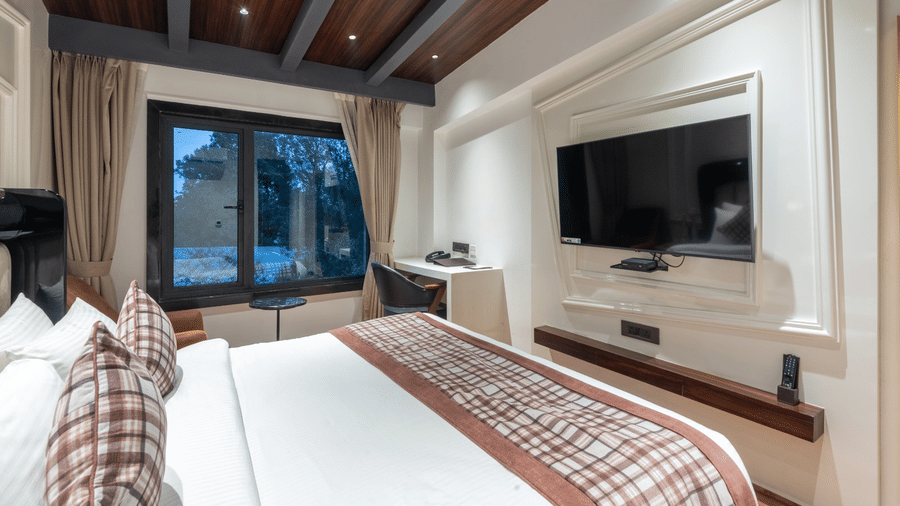 A hotel room with a bed, a wall-mounted flat-screen TV on a white panel, and a desk under a window | Lawrence Solitaire Resort, Nainital