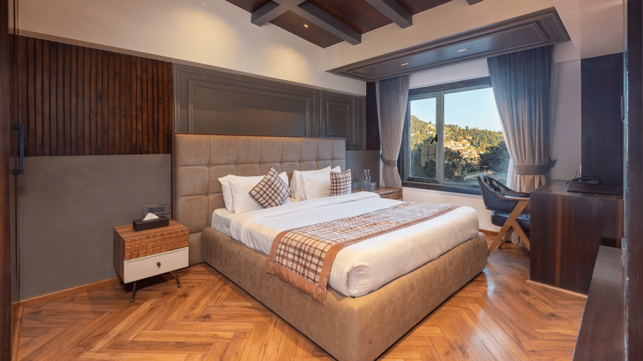 A hotel bedroom with a bed, small bedside tables, and a view of a forested hill through the window | Lawrence Solitaire Resort, Nainital