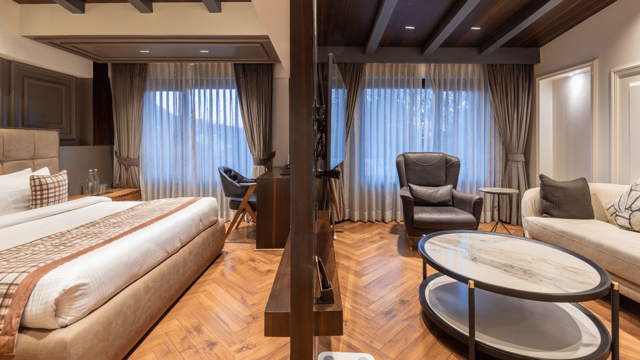 A hotel suite showing a large bed, a wooden room divider, a seating area, and windows with curtains | Lawrence Solitaire Resort, Nainital