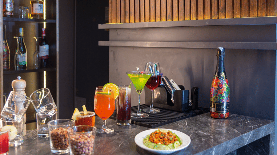 A bar counter featuring various colourful cocktails, glass bottles, and a plate of food against a wooden panelled wall | Lawrence Solitaire Resort, Nainital
