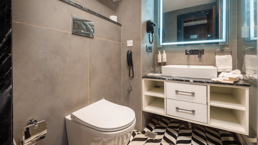 A bathroom with a wall-mounted toilet, a vanity with a sink, an illuminated mirror, and a chevron-patterned floor | Lawrence Solitaire Resort, Nainital