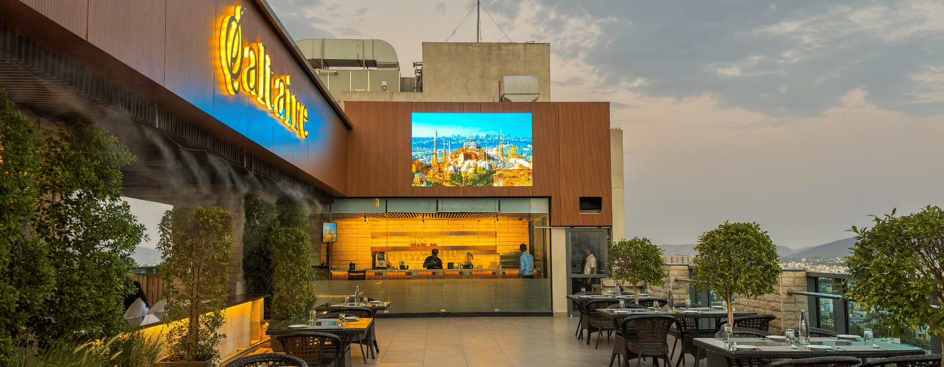 A view across the open terrace at the Saltaire restaurant at Pride Premier Udaipur. The area displays tables, chairs, potted plants, a bar counter towards the back, and a display screen mounted on a wall structure.