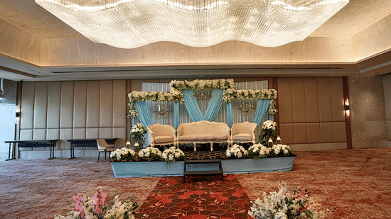 A stage set up for an event inside a hall, featuring seating, drapes, floral arrangements, a carpeted floor, and a chandelier.