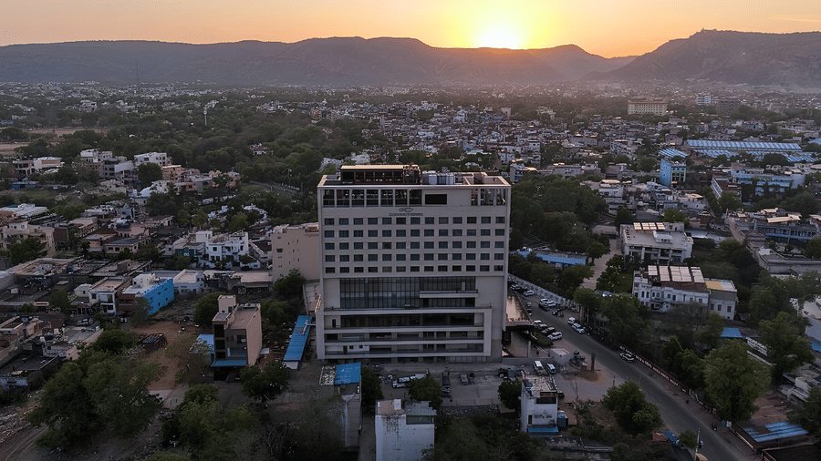 Aerial view of a hotel building surrounded by a town with trees and hills in the distance during sunset.