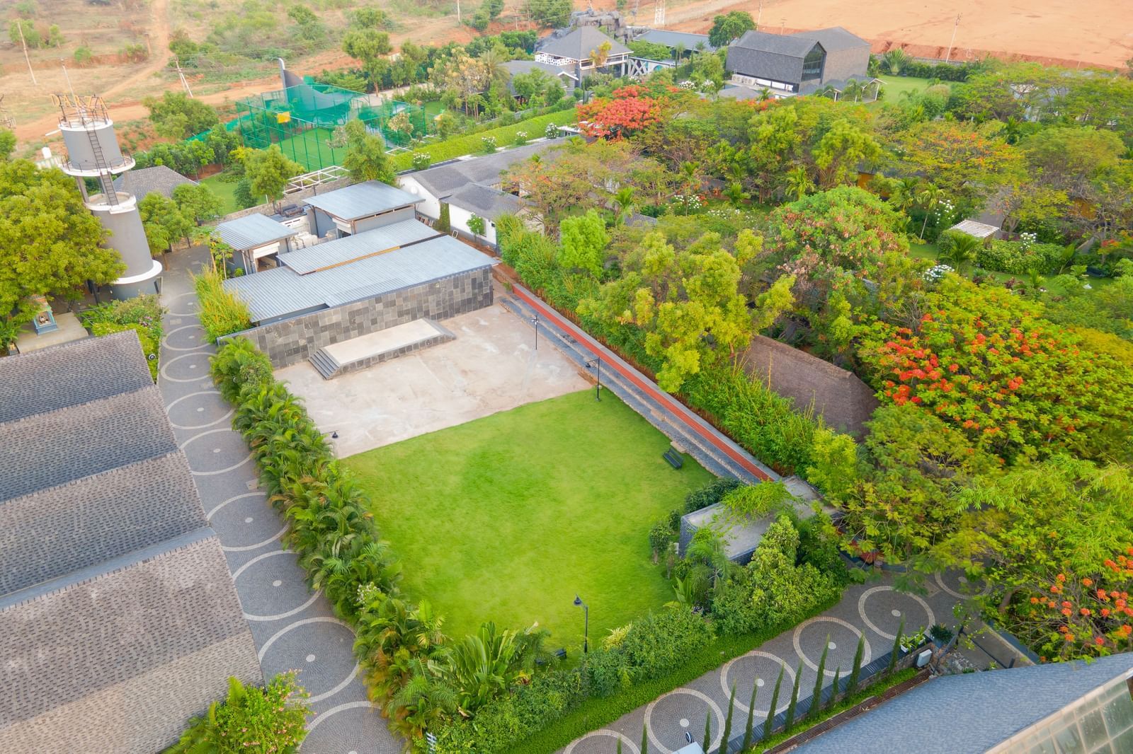 Enchante at Darza Resort featuring green lawn bordered by buildings, trees, and a pathway, part of a larger complex with varied landscaping under a bright sky.