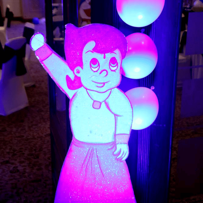 Kids party decoration at The Bristol Hotel, Gurgaon with an illuminated cartoon cutout beside a column of white balloons under purple and blue lighting.