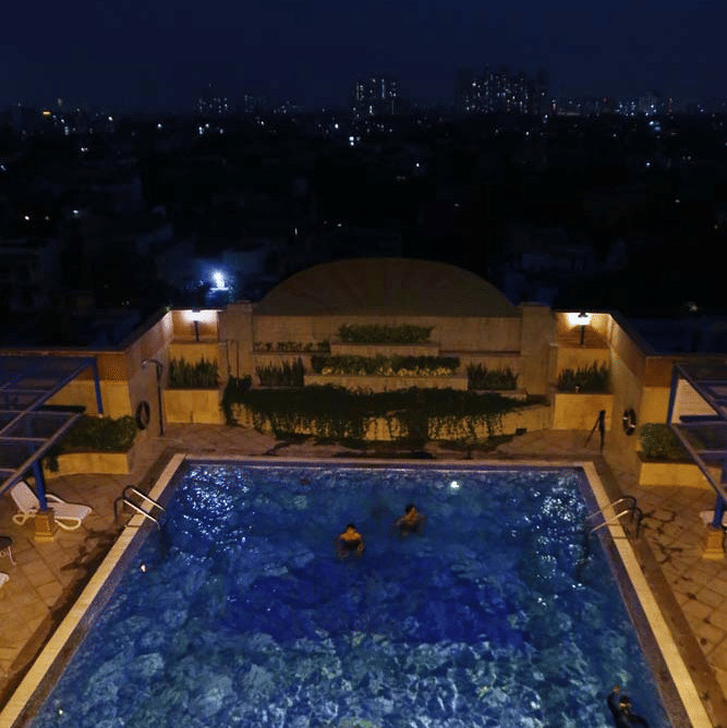 Rooftop swimming pool at The Bristol Hotel, Gurgaon at night with guests swimming and the illuminated city skyline visible in the background.